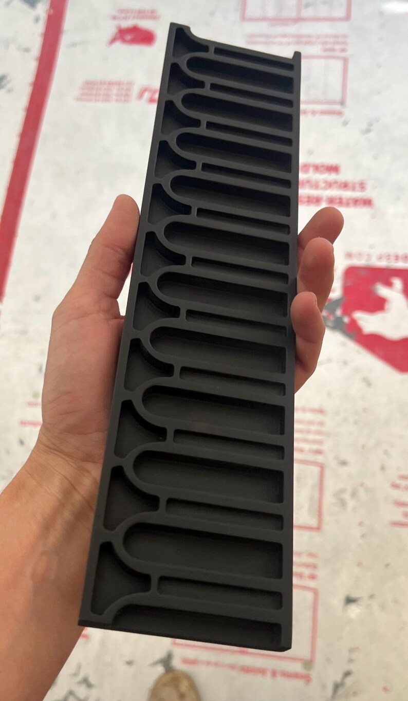 May include: A rectangular black plastic organizer tray featuring a series of uniform, elongated U-shaped slots arranged in a single column. The tray has a matte finish and a structured, rigid design with raised dividers between each individual compartment.