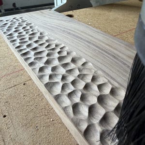 May include: A rectangular light brown wooden board featuring a textured surface with a repeating pattern of carved, rounded indentations along one side, positioned next to a black bristled brush tool on a workshop surface.