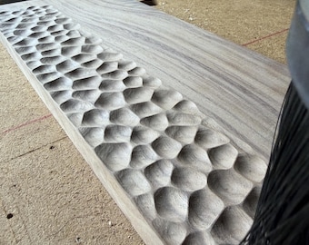 May include: A rectangular light brown wooden board featuring a textured surface with a repeating pattern of carved, rounded indentations along one side, positioned next to a black bristled brush tool on a workshop surface.