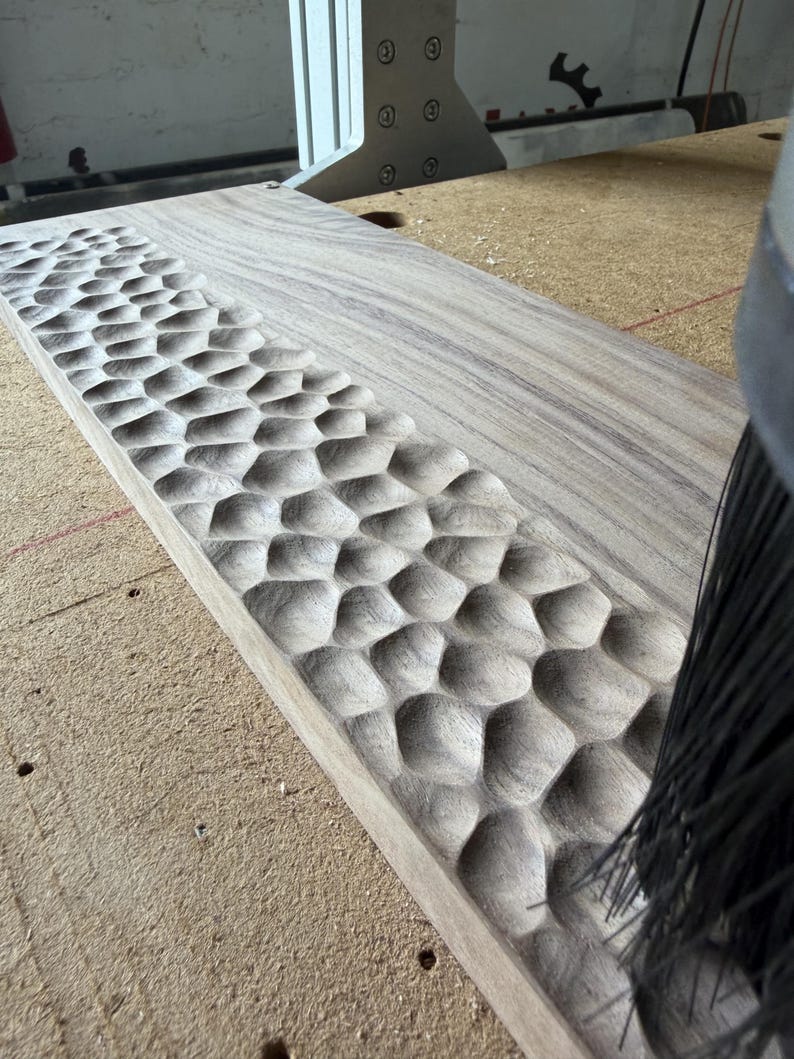 May include: A rectangular light brown wooden board featuring a textured surface with a repeating pattern of carved, rounded indentations along one side, positioned next to a black bristled brush tool on a workshop surface.
