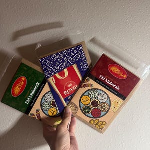 May include: Three Eid Mubarak greeting cards in clear plastic sleeves. The cards have various designs, including a green card with gold text, a red and gold card, and a card with a blue and white pattern and the word "Royal".