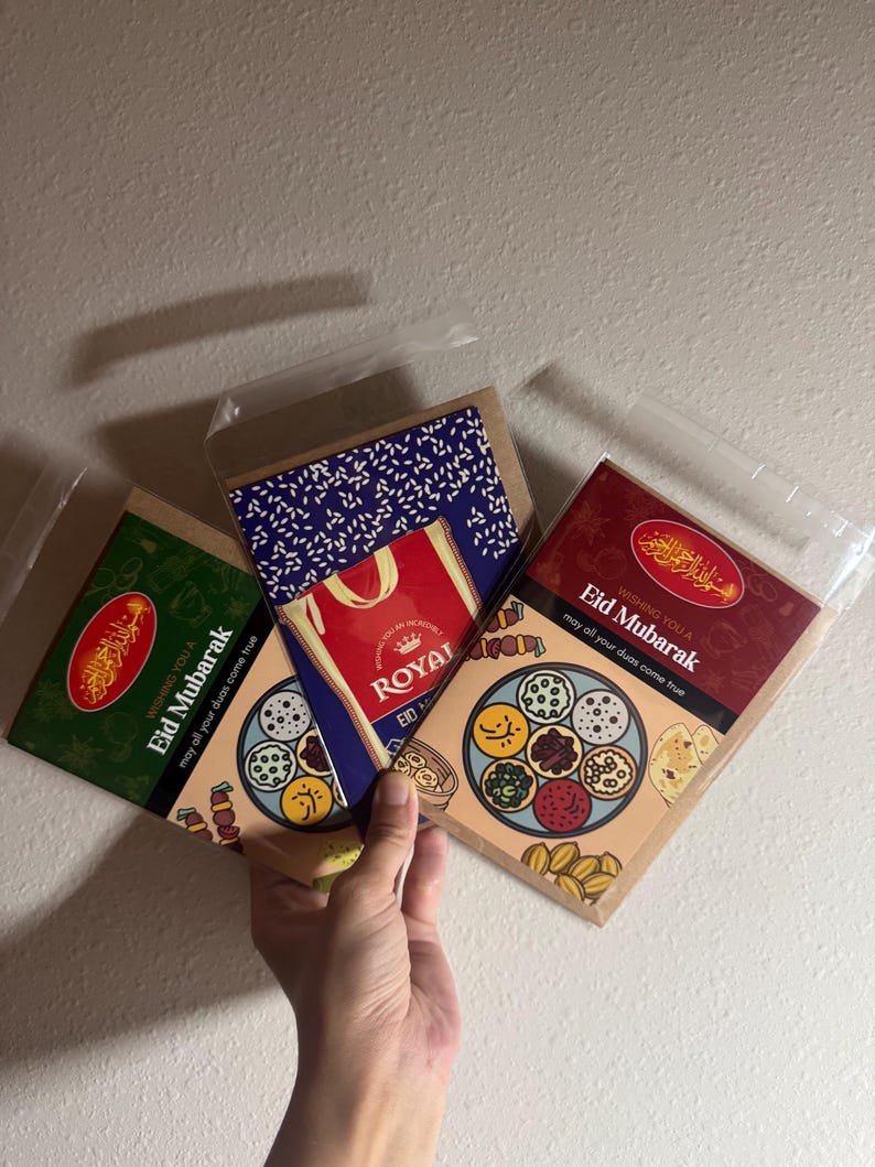 May include: Three Eid Mubarak greeting cards in clear plastic sleeves. The cards have various designs, including a green card with gold text, a red and gold card, and a card with a blue and white pattern and the word "Royal".