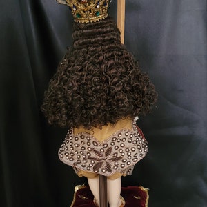 May include: A religious figurine featuring a gold crown adorned with colorful gemstones and a cross. The figurine has brown curly hair and is wearing a decorative garment. It stands on a detailed pedestal with red velvet shoes.