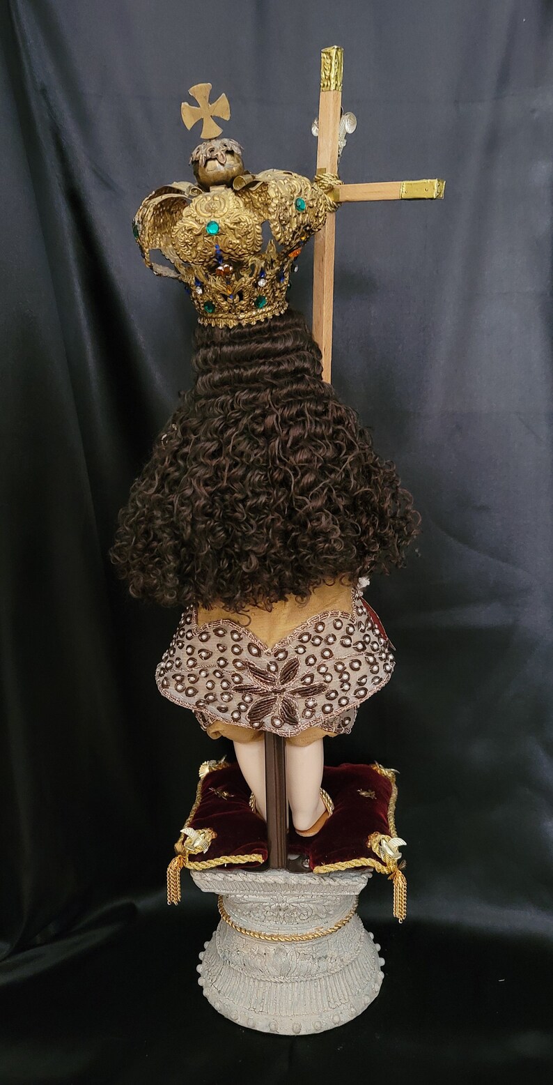 May include: A religious figurine featuring a gold crown adorned with colorful gemstones and a cross. The figurine has brown curly hair and is wearing a decorative garment. It stands on a detailed pedestal with red velvet shoes.