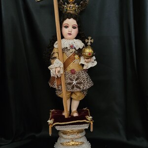 May include: A detailed doll of a young figure, adorned with a jeweled crown and holding a cross and orb. The doll wears a gold and brown outfit with intricate detailing, standing on a decorative pedestal. The background is a dark, draped fabric.