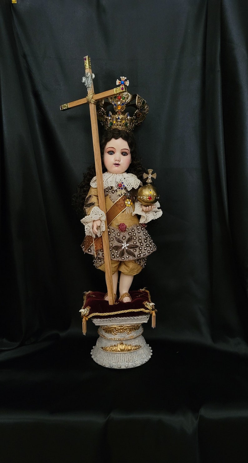 May include: A detailed doll of a young figure, adorned with a jeweled crown and holding a cross and orb. The doll wears a gold and brown outfit with intricate detailing, standing on a decorative pedestal. The background is a dark, draped fabric.