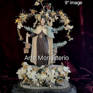May include: A religious statue featuring a woman in a dark robe and cream cape holding a child. The statue is surrounded by white flowers, gold accents, and a floral crown. The total height is 17 inches, with the image itself being 9 inches. The text "Arte Monasterio" is visible.