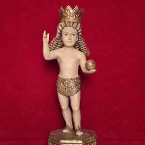 May include: A vintage statue of a young figure with a gold crown, holding a gold orb. The figure wears a gold patterned loincloth and stands on a gold decorated base. The statue has light skin and curly hair. The background is a solid red.