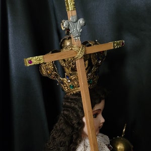May include: A detailed religious figurine of a child, wearing a gold crown adorned with jewels. The child holds a wooden cross with gold accents and a decorative silver emblem. The figurine is dressed in a gold and red outfit.