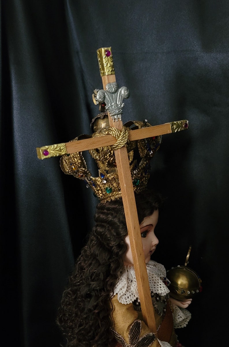 May include: A detailed religious figurine of a child, wearing a gold crown adorned with jewels. The child holds a wooden cross with gold accents and a decorative silver emblem. The figurine is dressed in a gold and red outfit.