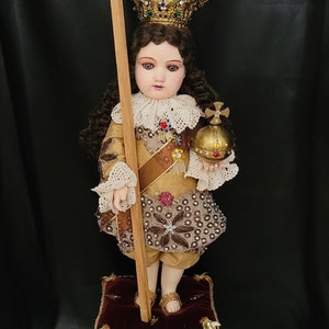 May include: A detailed doll of a child in regal attire, holding a wooden cross and a golden orb. The doll wears a jeweled crown, a gold and brown outfit with intricate embroidery, and stands on a decorative pedestal.