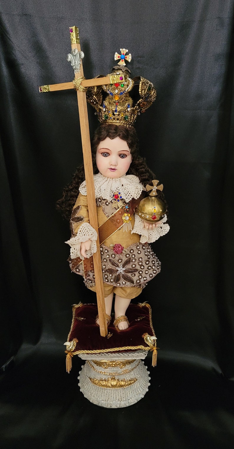 May include: A detailed doll of a child in regal attire, holding a wooden cross and a golden orb. The doll wears a jeweled crown, a gold and brown outfit with intricate embroidery, and stands on a decorative pedestal.