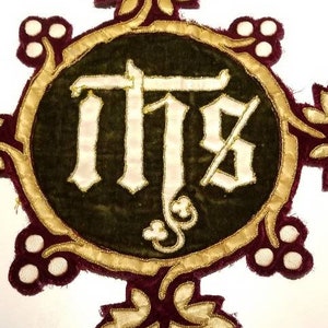 May include: A circular embroidered patch with a gold border and burgundy velvet background. The center features a white embroidered monogram "IHS" with gold outlining. The patch is decorated with gold and white embroidered floral motifs.