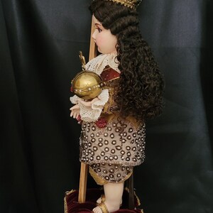 May include: A detailed religious figurine of a child, adorned with a gold crown and holding a golden orb. The figure wears an ornate outfit with intricate beadwork and stands on a decorative pedestal. A wooden cross is behind the figure.