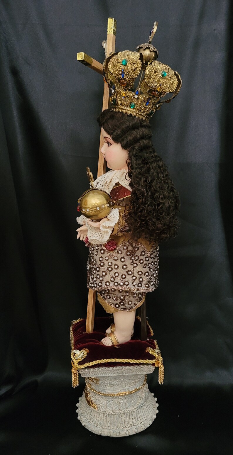 May include: A detailed religious figurine of a child, adorned with a gold crown and holding a golden orb. The figure wears an ornate outfit with intricate beadwork and stands on a decorative pedestal. A wooden cross is behind the figure.