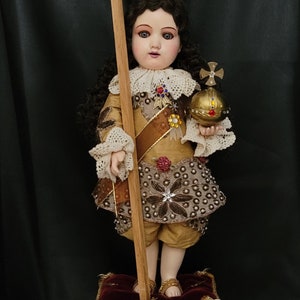 May include: A detailed doll of a young figure, adorned with a jeweled crown and holding a cross and orb. The doll is dressed in gold and brown clothing, standing on a decorative pedestal. Total height is 25 inches, image height is 14.5 inches.