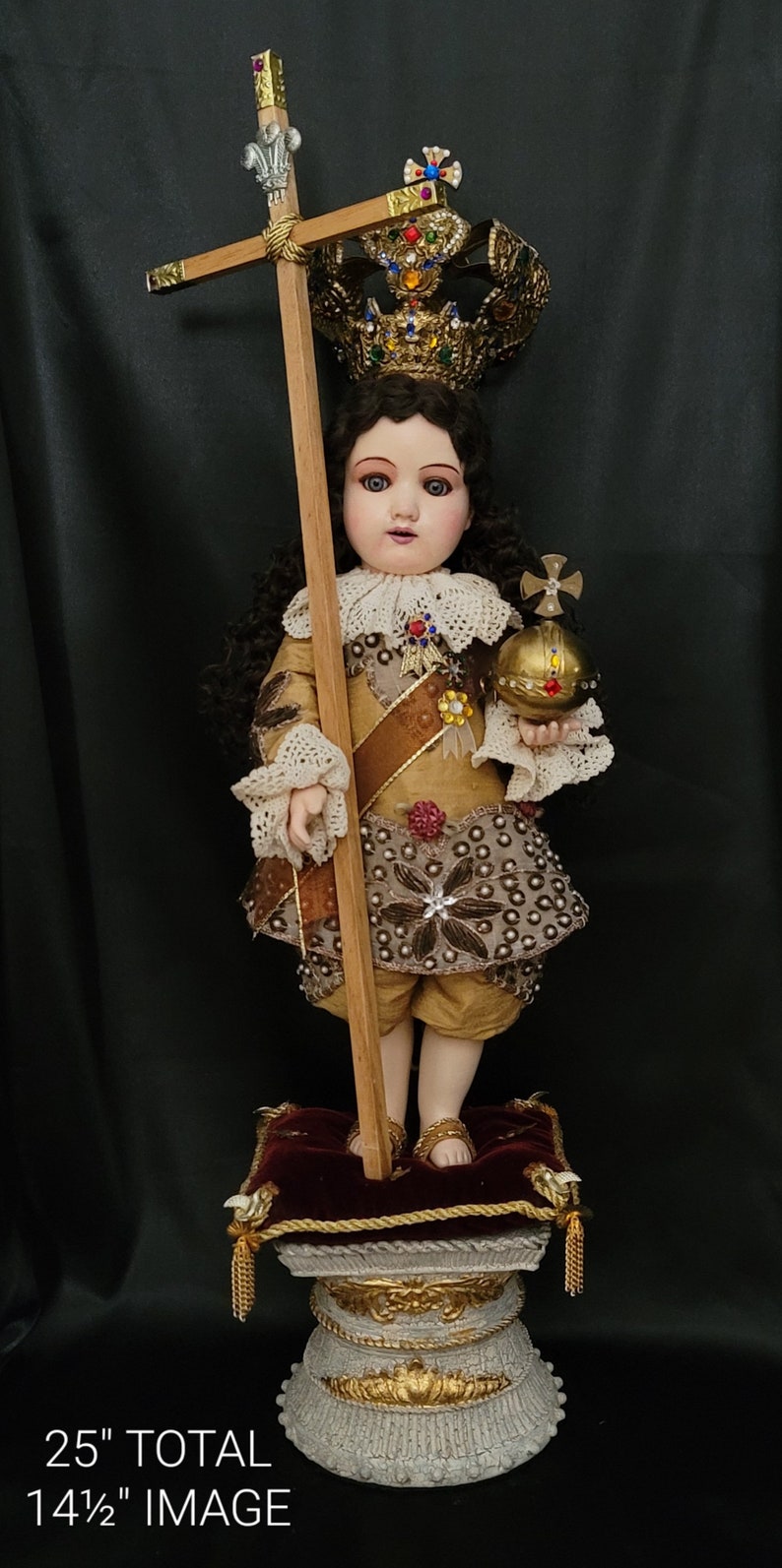 May include: A detailed doll of a young figure, adorned with a jeweled crown and holding a cross and orb. The doll is dressed in gold and brown clothing, standing on a decorative pedestal. Total height is 25 inches, image height is 14.5 inches.