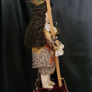 May include: A detailed side view of a religious figurine. The figurine wears a gold crown, ornate clothing, and holds a cross. It stands on a decorative pedestal with a red cushion. The figurine has long, curly brown hair.