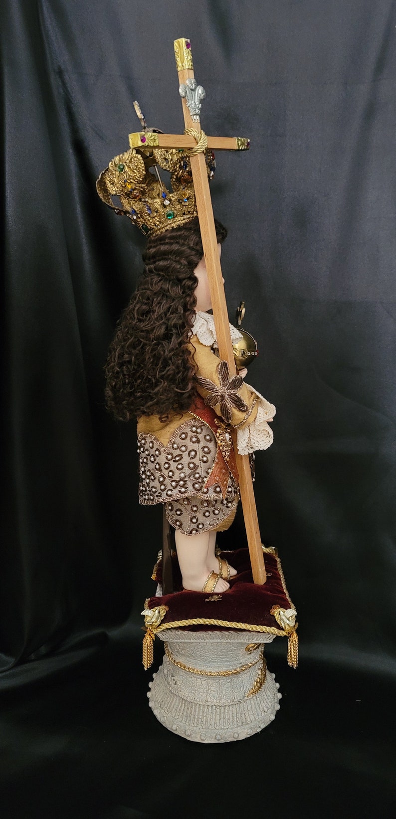 May include: A detailed side view of a religious figurine. The figurine wears a gold crown, ornate clothing, and holds a cross. It stands on a decorative pedestal with a red cushion. The figurine has long, curly brown hair.