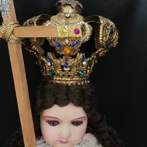 May include: A detailed doll with a golden crown adorned with colorful jewels. The doll has brown hair, blue eyes, and is wearing a gold and red outfit. It holds a wooden staff with a decorative top. The background is black.
