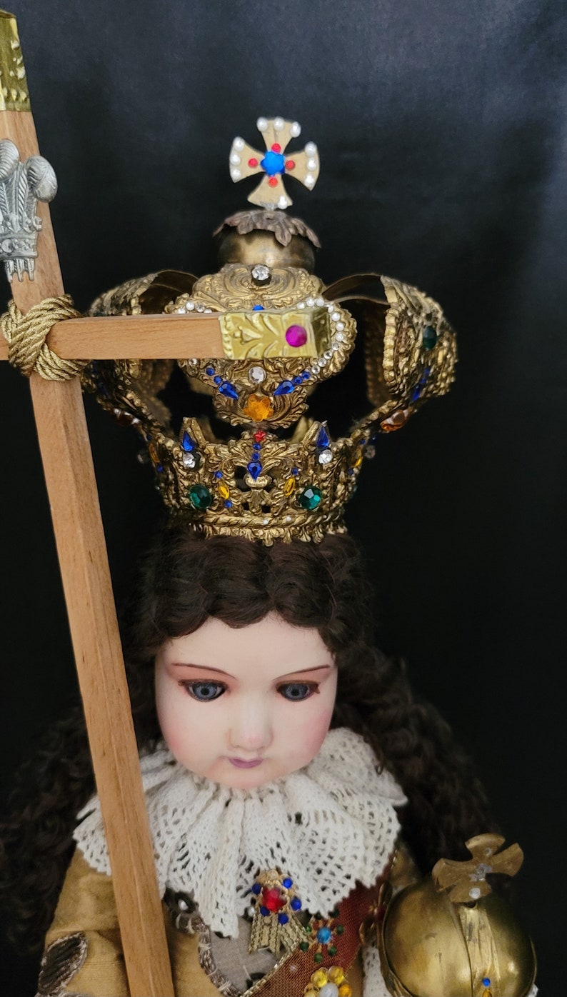 May include: A detailed doll with a golden crown adorned with colorful jewels. The doll has brown hair, blue eyes, and is wearing a gold and red outfit. It holds a wooden staff with a decorative top. The background is black.