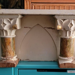 May include: A white wooden shelf with two decorative columns on either side. The columns are painted with gold and green accents. The shelf has a pointed arch in the center.