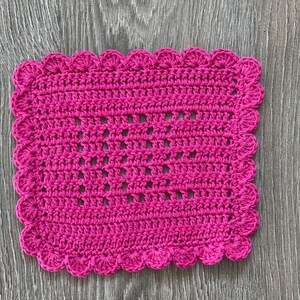 May include: A pink crocheted square with a scalloped edge. The crochet is a delicate pattern with open spaces.