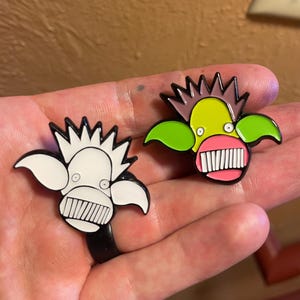 May include: Two enamel pins featuring cartoon monster faces. The first pin is white with black outlines and a pink mouth with white teeth. The second pin is green with black outlines, a pink mouth with white teeth, and a pink nose. Both pins have spiky hair.