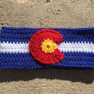 May include: Crocheted headband with the Colorado state flag design. The headband is blue with a white stripe and a red circle with a yellow center.