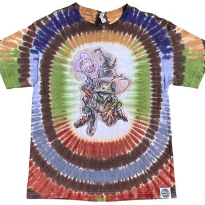 May include: A tie-dye t-shirt with a circular pattern in shades of brown, orange, green, blue, and red. The shirt features a graphic of a frog wearing a hat and holding a staff. The shirt has a tag at the neckline and a small logo at the bottom.