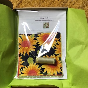 May include: A craft kit containing sunflower fabric, thread, and instructions. The fabric displays large yellow sunflowers on a dark blue backdrop. The kit includes a spool of beige thread and instructions for patchwork squares.