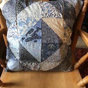 Op de afbeelding: Een blauw en beige patchwork kussen met een geometrisch patroon. Het kussen ligt op een houten stoel.