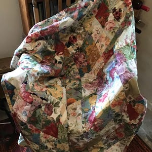 May include: A floral quilt with a diamond pattern in shades of pink, red, green, and yellow. The quilt is draped over a wooden chair.