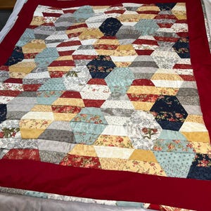 May include: A red, white and blue quilt with a geometric pattern. The quilt is made of various fabrics with floral and geometric designs. The quilt is bordered with a red fabric.