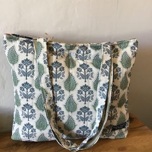 Op de afbeelding: Een gewatteerde tote bag met een herhalend patroon van blauwe en groene bloem- en bladmotieven op een gebroken witte achtergrond. De tas heeft twee schouderbanden en een klein zwart rechthoekig label met witte tekst Cotton Crafts by Rinda.