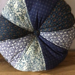 May include: A round, tufted cushion with a patchwork design. The cushion is made of different fabrics with floral patterns in shades of blue, green, and white. The cushion has a button in the centre.
