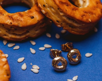 May include: Two gold-toned earrings shaped like a bagel on a blue background with white sesame seeds scattered around them.