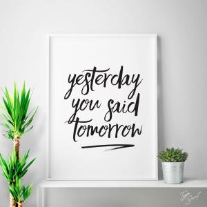 Op de afbeelding: Witgelijste kunstdruk met de tekst "yesterday you said tomorrow" in zwart script. De print wordt weergegeven op een witte plank met een kleine vetplant in pot en een hoge groene plant. Het thema is motiverend.