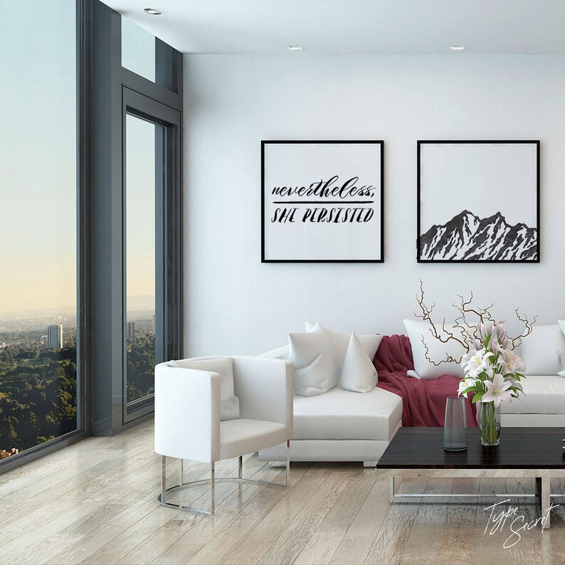 May include: A white living room with a white couch, a white chair, and two black framed prints on the wall. The print on the left says "nevertheless, she persisted". The print on the right shows a black and white mountain range.
