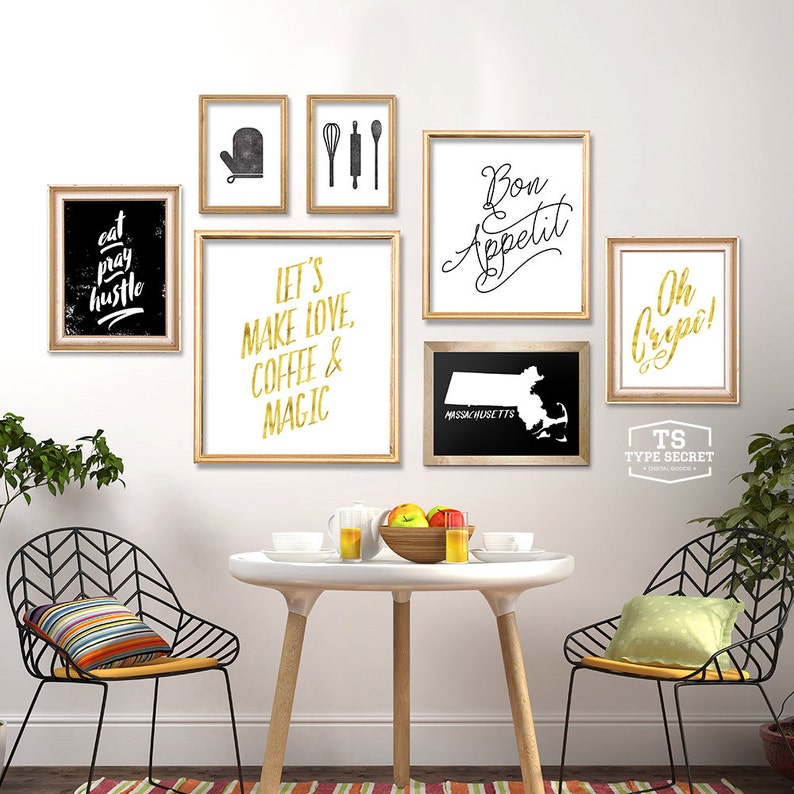 May include: A white wall with a gallery wall of framed prints. The prints feature black and gold text and graphics. The prints include the text "Eat Pray Hustle", "Let's Make Love, Coffee & Magic", "Bon Appetit", "Oh Cripe!" and "Massachusetts". There are two black metal chairs with yellow cushions and a white table with a bowl of fruit and glasses on it.