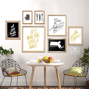 May include: A white wall with a gallery wall of framed prints. The prints feature black and gold text and graphics. The prints include the text "Eat Pray Hustle", "Let's Make Love, Coffee & Magic", "Bon Appetit", "Oh Cripe!" and "Massachusetts". There are two black metal chairs with yellow cushions and a white table with a bowl of fruit and glasses on it.