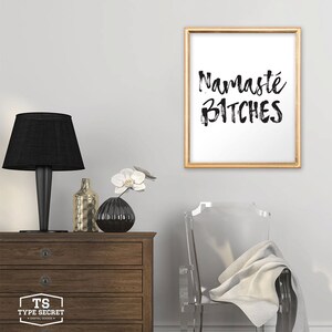 Puede incluir: Una impresi&oacute;n enmarcada en dorado con el texto "Namaste Bitches" en escritura de pincel negro sobre fondo blanco. La impresi&oacute;n est&aacute; colgada en una pared blanca sobre una c&oacute;moda de madera con una l&aacute;mpara negra y un jarr&oacute;n con flores blancas.