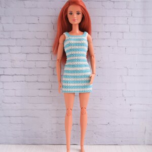 May include: A doll wearing a blue and white striped crochet dress. The doll has long red hair and is standing in front of a white brick wall.