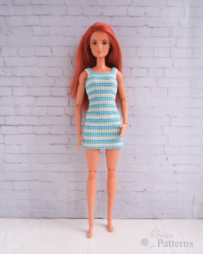 May include: A doll wearing a blue and white striped crochet dress. The doll has long red hair and is standing in front of a white brick wall.