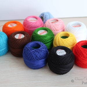 Op de afbeelding: Een verzameling van 12 bollen borduurgaren in verschillende kleuren, waaronder blauw, groen, rood, oranje, geel, bruin, roze, wit en zwart. De bollen zijn in een cirkel op een houten oppervlak gerangschikt.