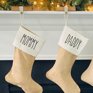 May include: Two Christmas stockings with burlap bodies and white tops. The word "MOMMY" is printed on one stocking, and "DADDY" is printed on the other in black script. The stockings hang from a white mantelpiece decorated with greenery and lights.