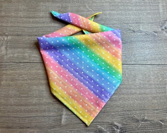 May include: A colorful rainbow-striped bandana with white polka dots. The fabric is a blend of yellow, orange, pink, purple, and blue. The bandana is tied at the top, creating a triangular shape. This accessory is suitable for pets.