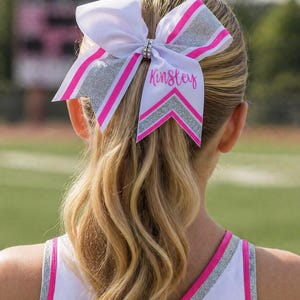May include: A white cheerleading bow with pink and silver accents, featuring the name "Kinsley" in cursive. The bow is adorned with a rhinestone center and is worn in a ponytail. The cheerleader's uniform is white and pink.