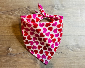May include: A white dog bandana with a pattern of red and pink hearts. The bandana is tied at the top, creating a triangle shape. The fabric is soft and likely cotton.