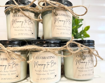 May include: A stack of small glass jars with black lids, filled with white wax candles. Each jar is adorned with a label that reads "Celebrating Mommy to Bee" and the date of a baby shower. The jars are tied with twine and a bee charm.